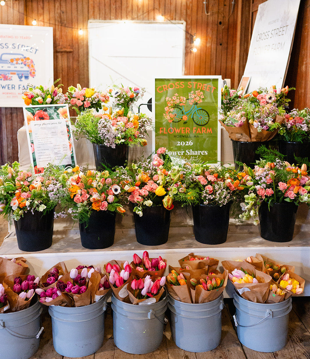 Floral arrangements and flower shares displayed on a table with wooden walls and string lights.