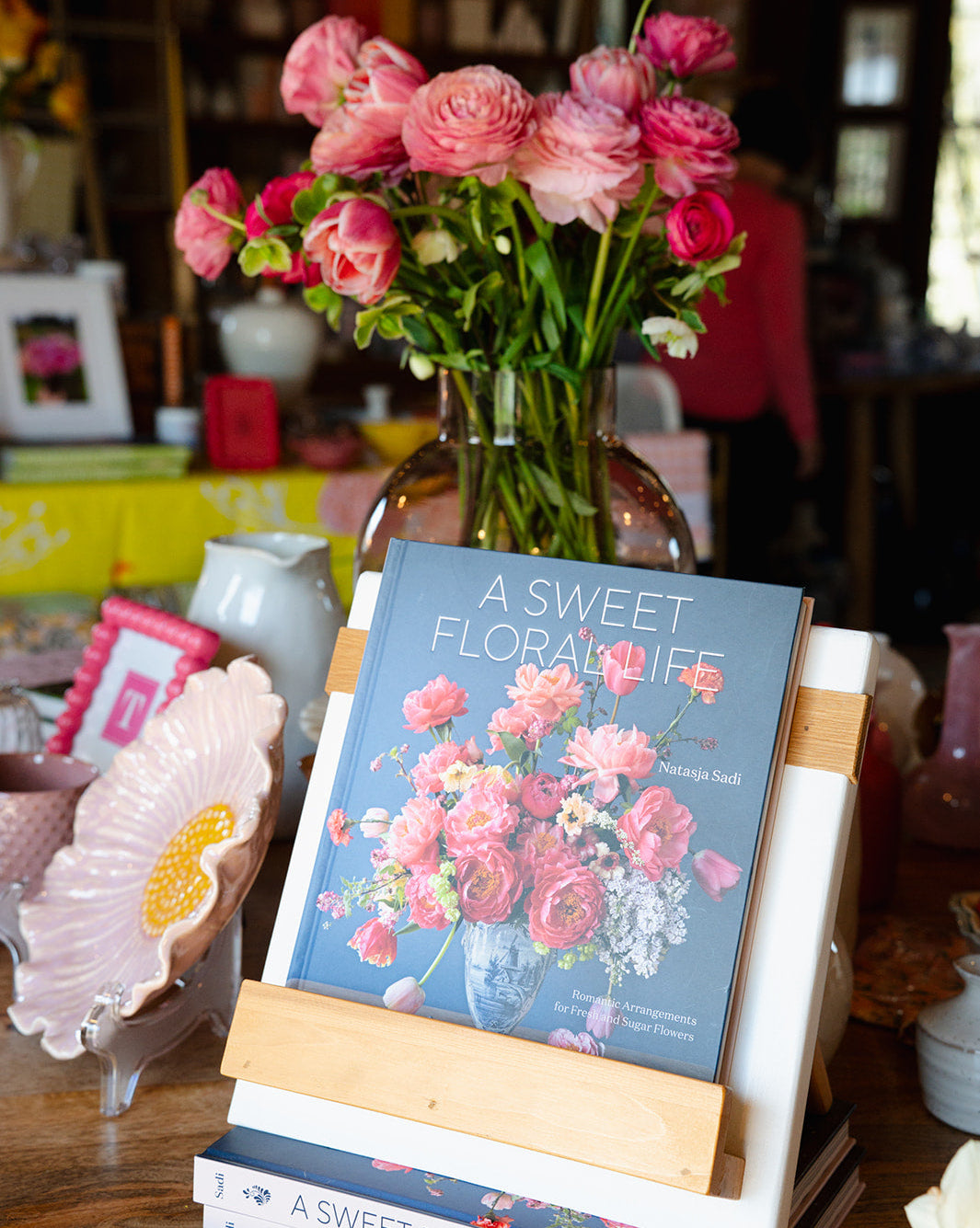 Book titled 'A Sweet Floral Life' on a wooden stand with a vase of pink flowers in the background.